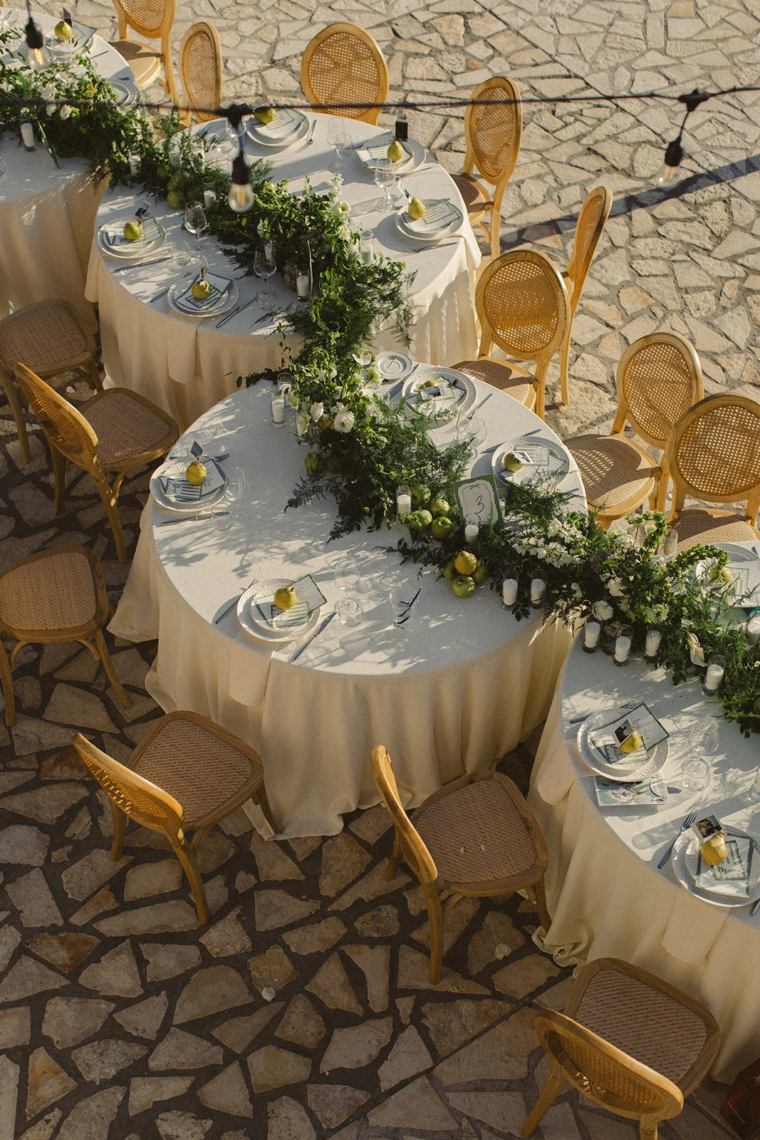 Flottie wedding decor and organisation: overhead view of outdoor round wedding tables styled with lush green garlands, white flowers, and fresh pears as part of the event decor