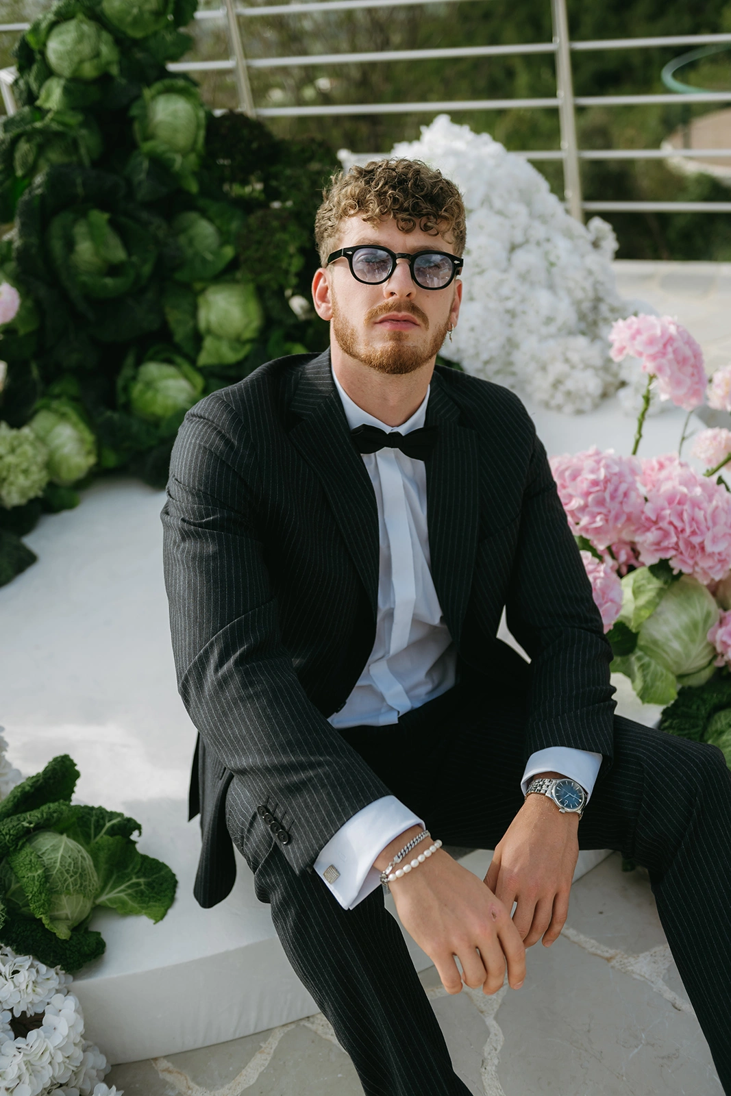 Flottie The Cabbage Affair wedding decor and organisation: stylish groom in a pinstripe suit and bow tie posing thoughtfully on the white stage decorated with fresh cabbages and floral elements