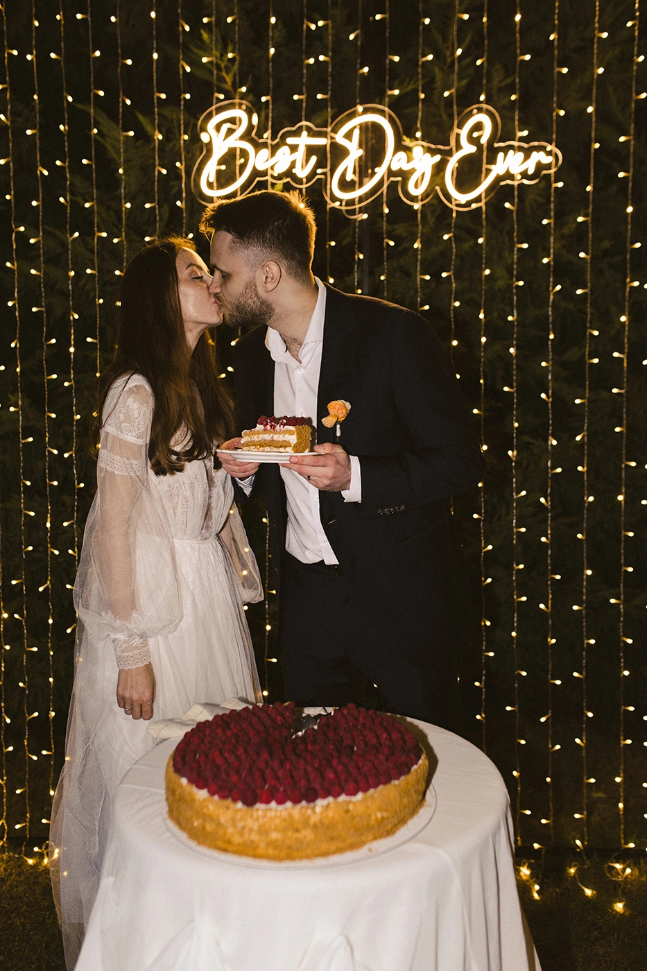 Flottie Peach Haze wedding decor and organisation: bride and groom kissing in front of a warm string light backdrop and "Best Day Ever" neon sign with a berry cake