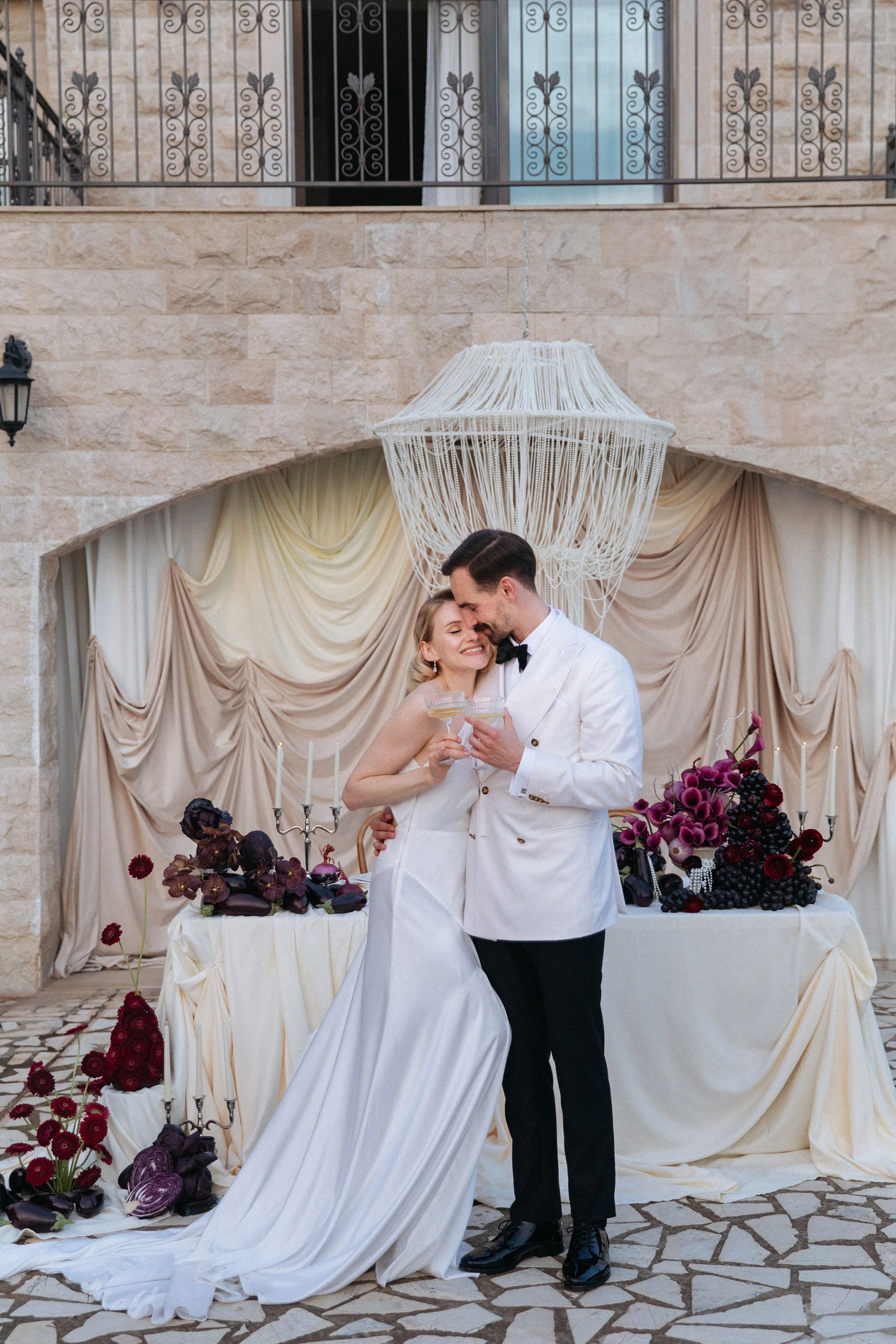 Flottie Shades of Burgundy wedding decor and organisation: bride in a minimalist white gown and groom in a white double-breasted jacket toasting with champagne in front of a draped arch