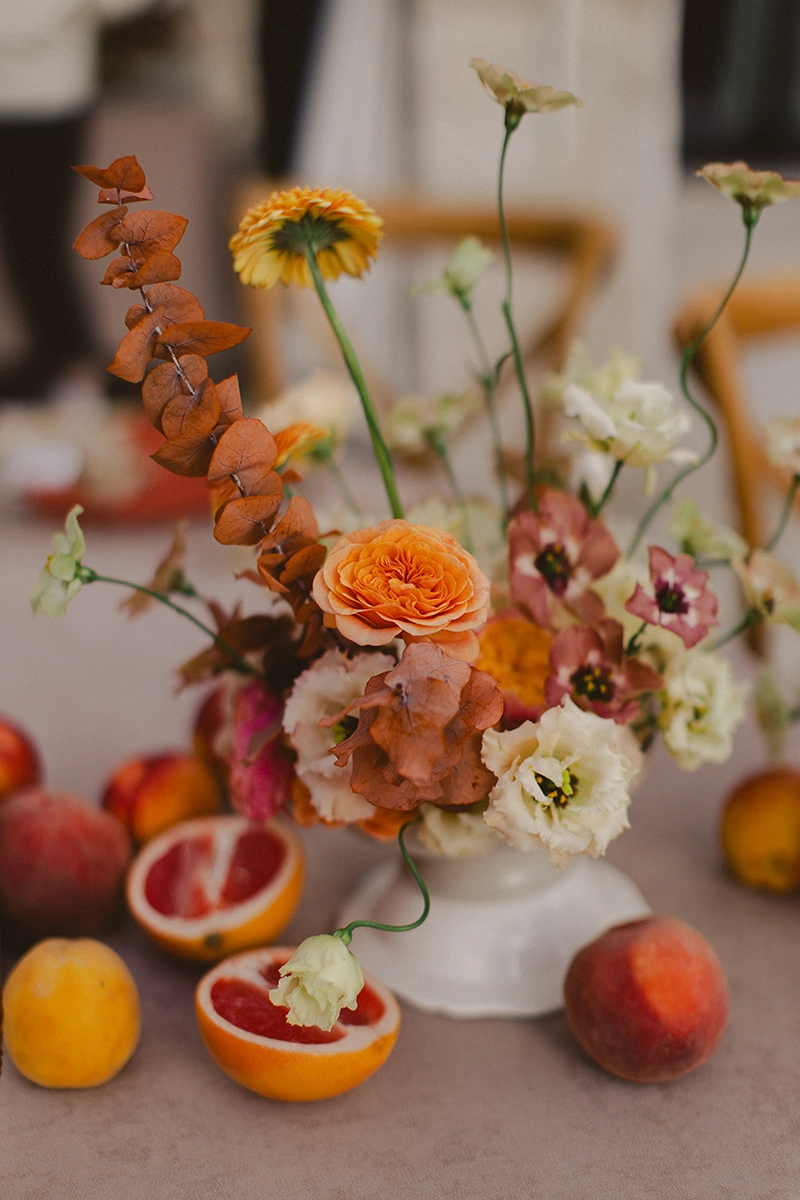 Flottie Peach Haze wedding decor and organisation: close-up of a delicate floral centerpiece arrangement with pastel roses and fresh peaches on a table