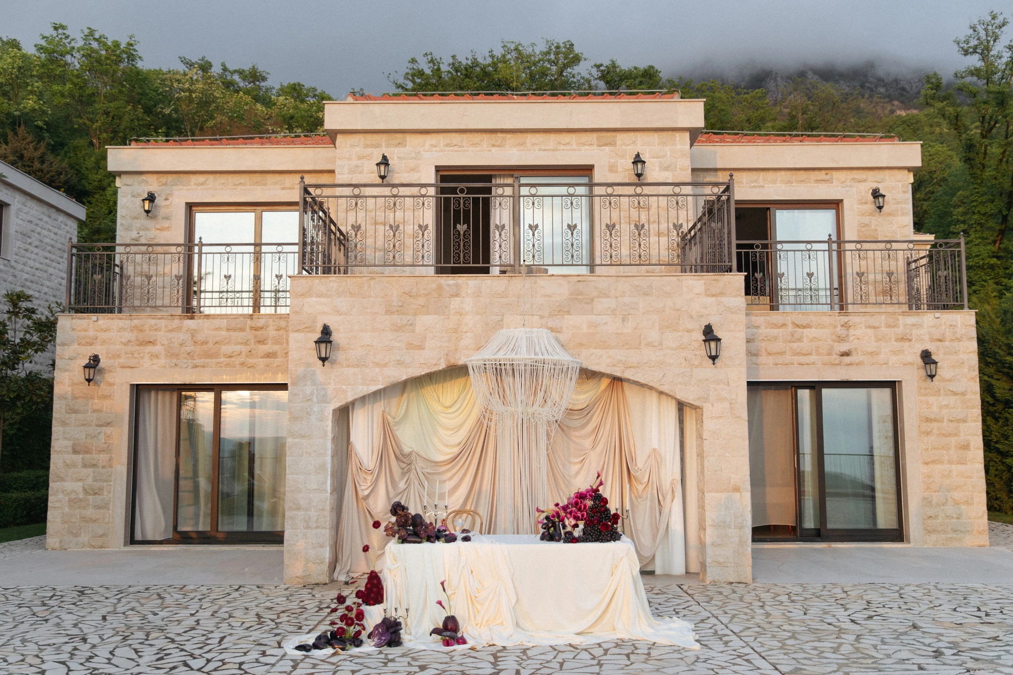 Flottie Shades of Burgundy wedding decor and organisation: exterior view of a stone villa featuring a draped ceremony backdrop, ornate chandelier, and a styled sweetheart table adorned with burgundy florals