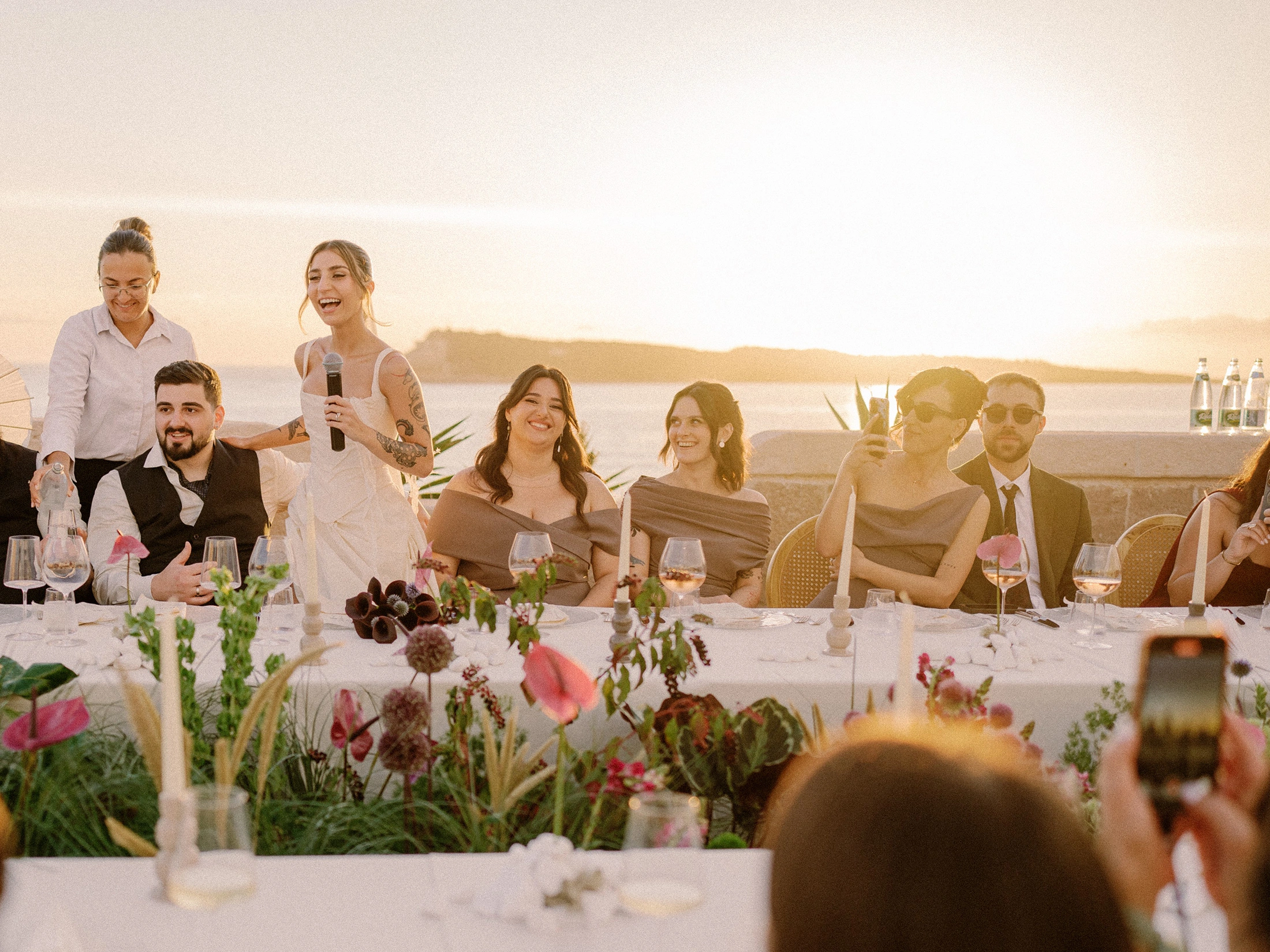 Flottie wedding decor and organisation: joyful bride speaking into a microphone during a wedding reception dinner with guests, candlelit tablescape, and floral arrangements
