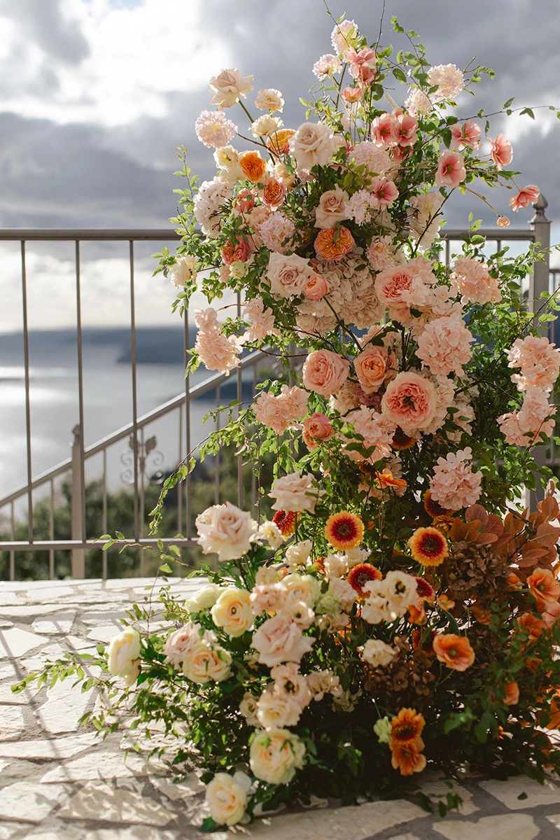Flottie Peach Haze wedding decor and organisation: lush peach and pink floral arrangement column set against a scenic outdoor coastal background