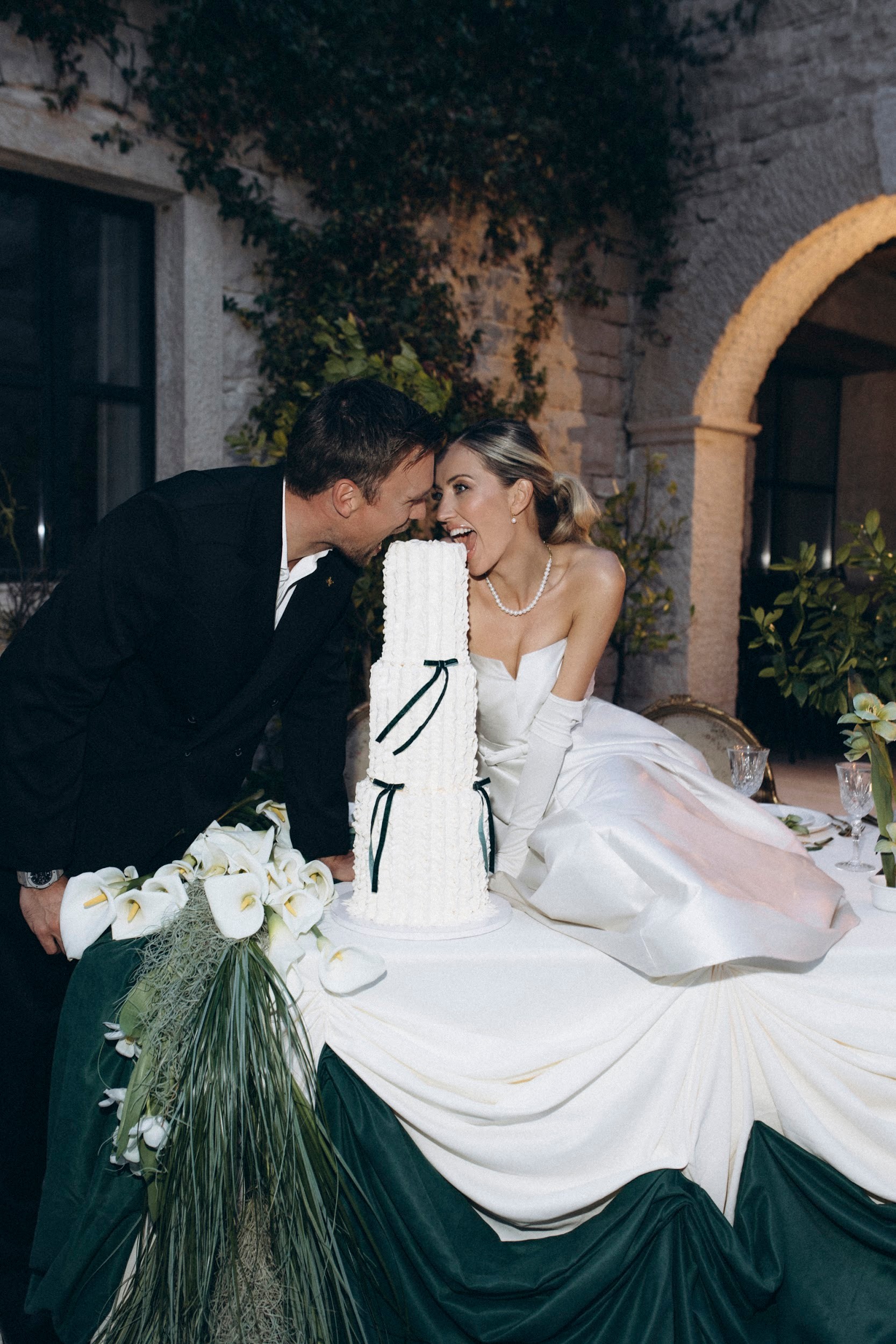 Flottie flower decor, wedding, and organisation at Borgo Lapis in Croatia: bride and groom playfully cutting a tiered wedding cake adorned with white calla lilies and rich green drapery