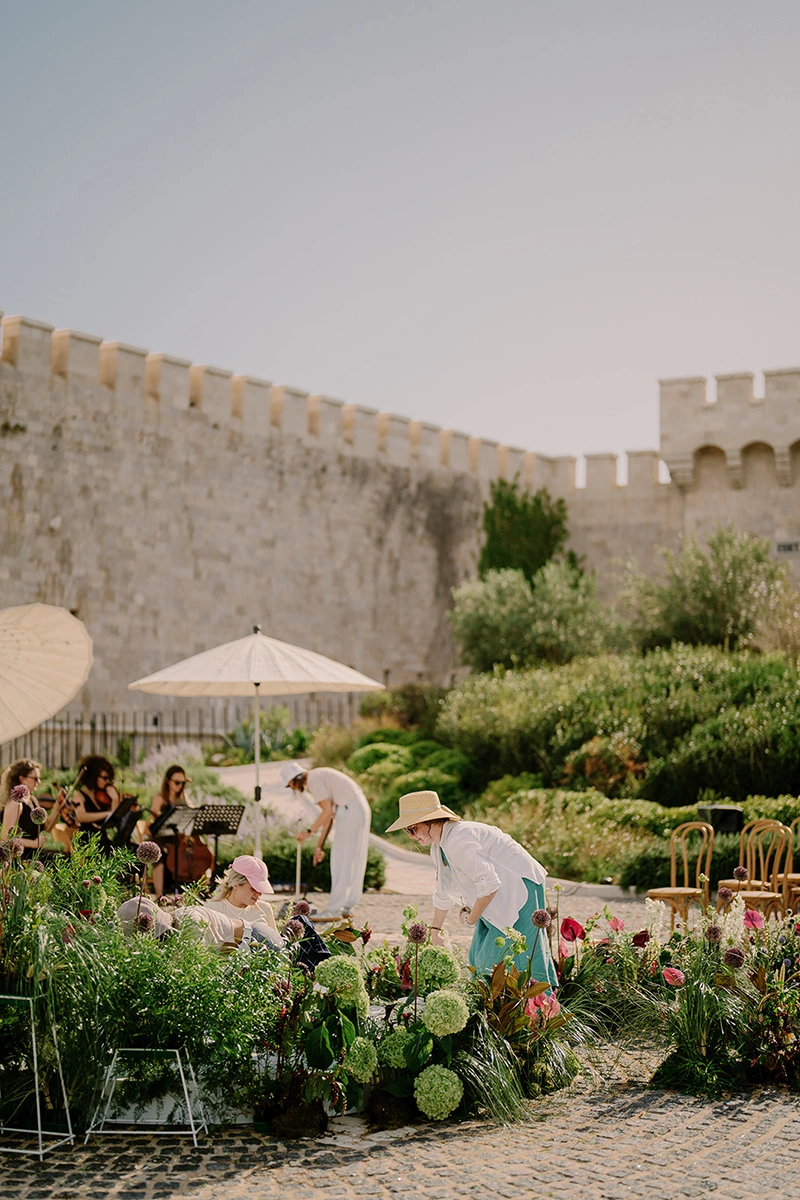 Flottie wedding decor and organisation: behind-the-scenes setup in an outdoor courtyard near a historic stone fortress wall, featuring floral designers arranging botanical installations and event elements