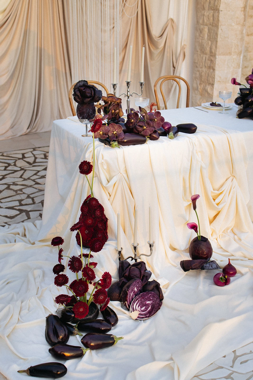 Flottie Shades of Burgundy wedding decor and organisation: close-up of a dramatic tablescape arrangement featuring vibrant red gerberas, fresh eggplants, sliced red cabbage, and a silver candelabra