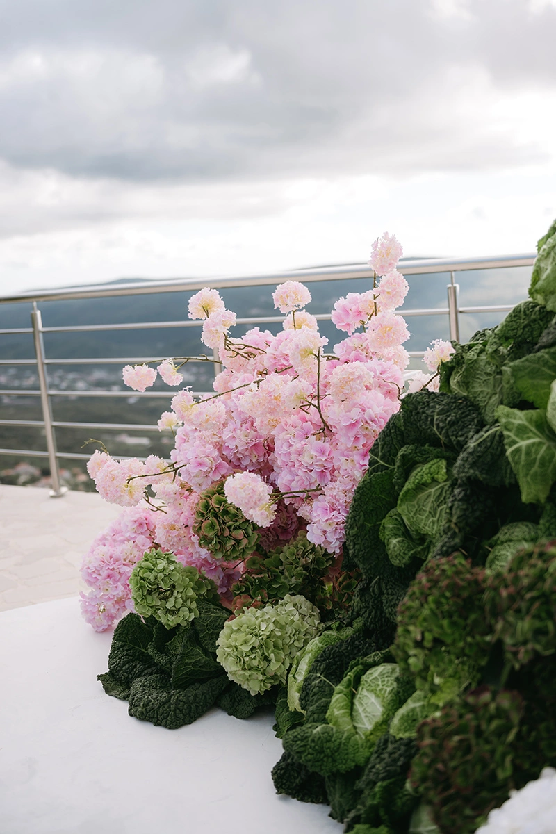 Flottie The Cabbage Affair wedding decor and organisation: modern ceremony setup featuring transparent ghost chairs and a unique sculptural altar installation made of fresh cabbages and pink and white florals overlooking a mountain view