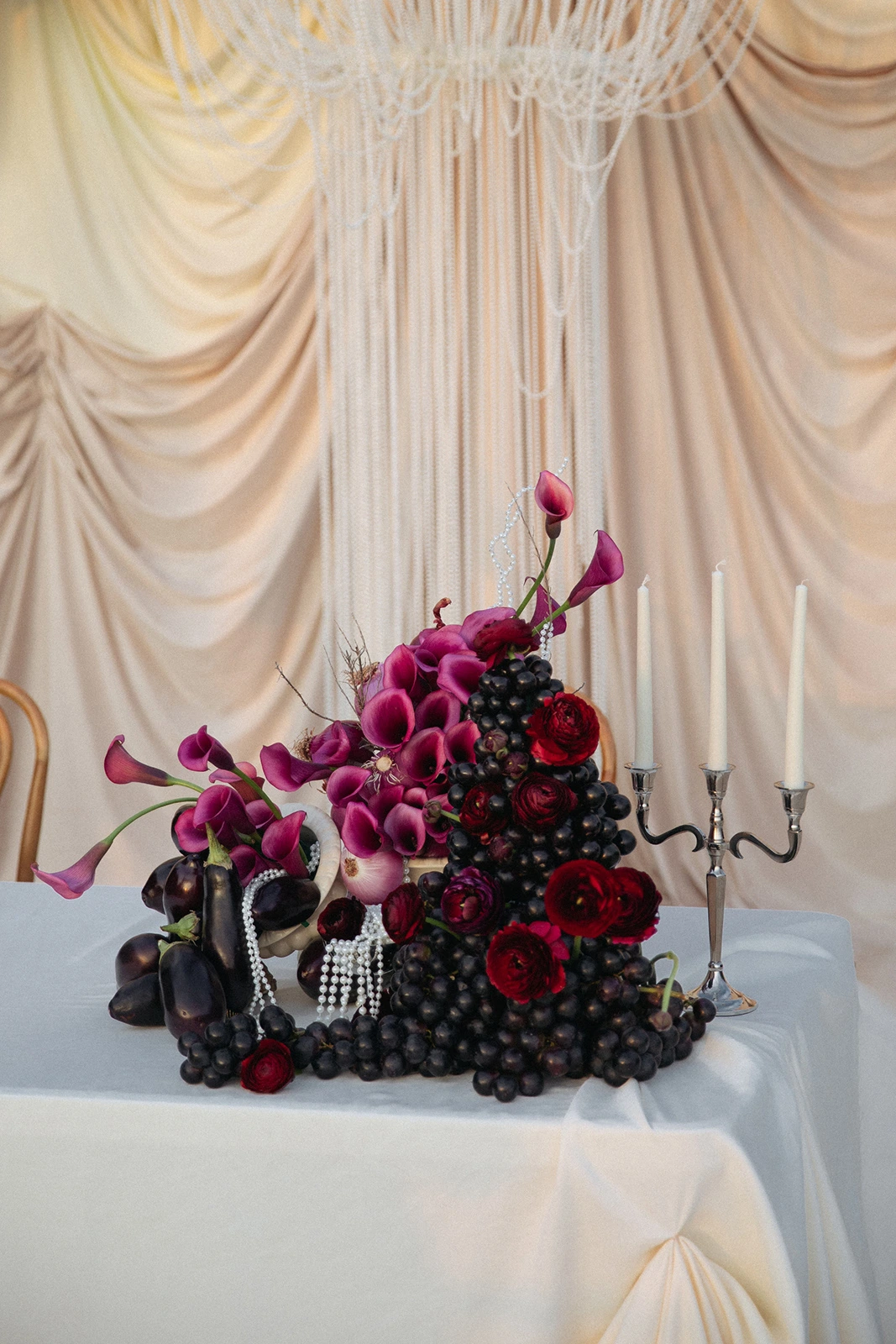 Flottie Shades of Burgundy wedding decor and organisation: close-up of a dramatic tablescape arrangement featuring vibrant red gerberas, fresh eggplants, sliced red cabbage, and a silver candelabra