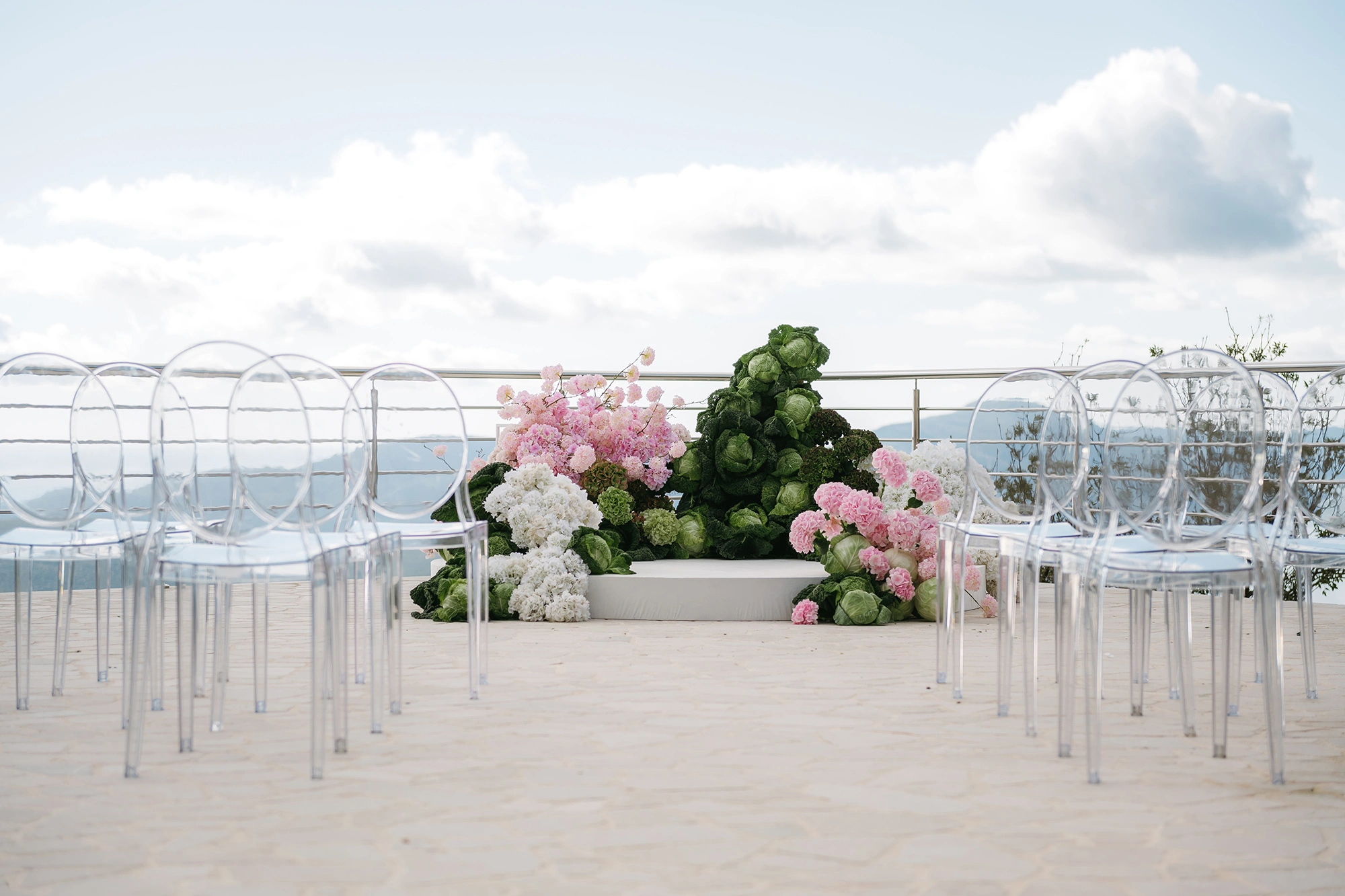 Flottie The Cabbage Affair wedding decor and organisation: modern ceremony setup featuring transparent ghost chairs and a unique sculptural altar installation made of fresh cabbages and pink and white florals overlooking a mountain view