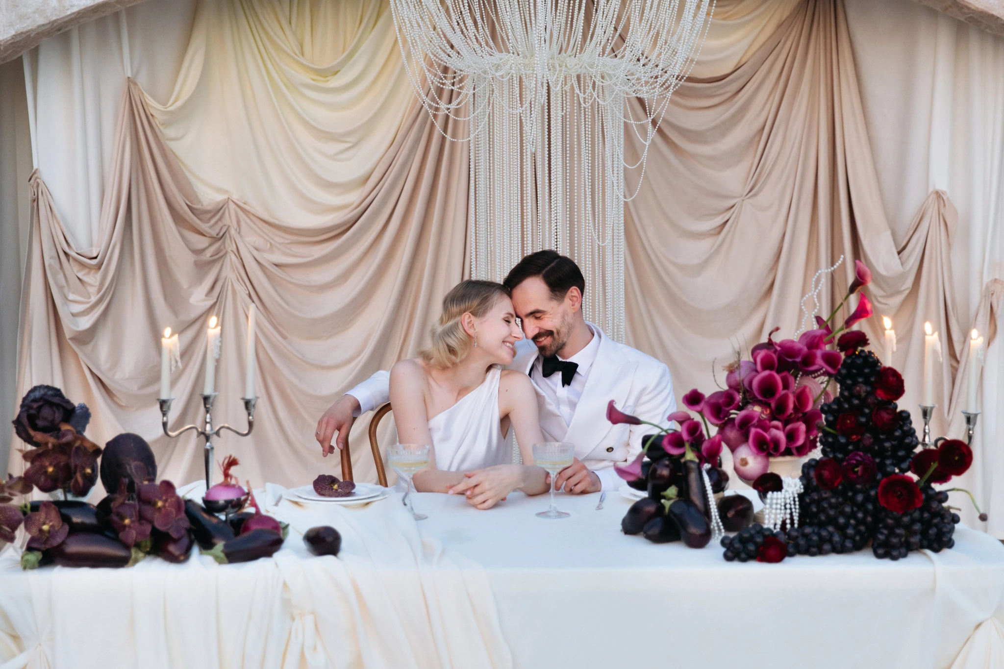 Flottie Shades of Burgundy wedding decor and organisation: bride in a minimalist white gown and groom in a white double-breasted jacket toasting with champagne in front of a draped arch