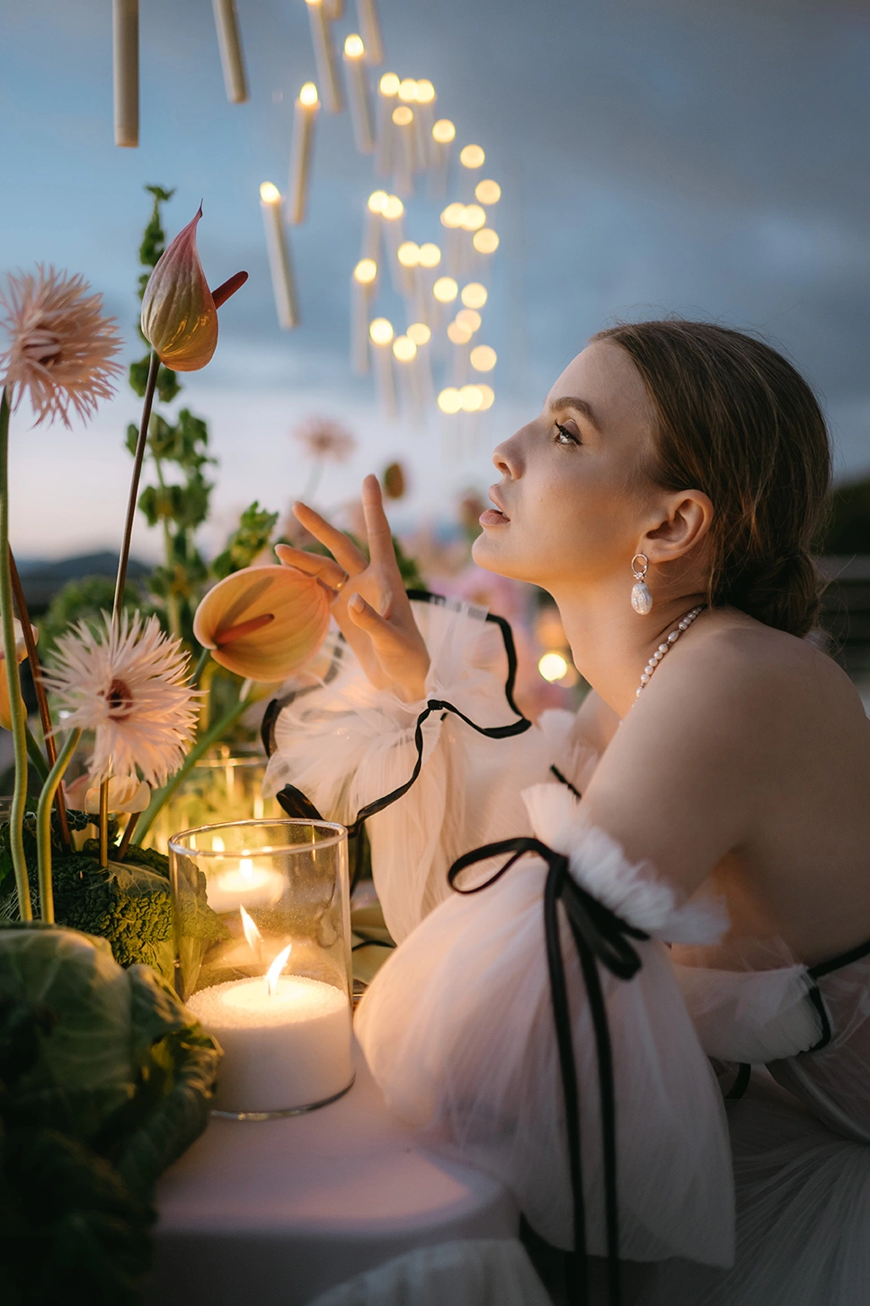 Flottie The Cabbage Affair wedding decor and organisation: close-up portrait of a bride in a tulle dress with black ribbons gently touching exotic anthurium flowers and gerberas at a candlelit evening reception table