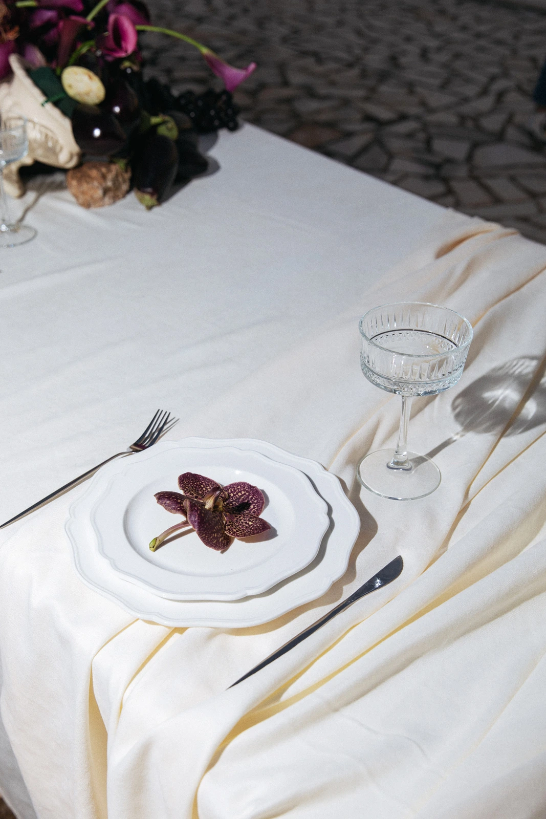 Flottie Shades of Burgundy wedding decor and organisation: elegant table setting with layered white plates, a dark purple orchid, a crystal coupe glass, and silverware on a draped linen tablecloth