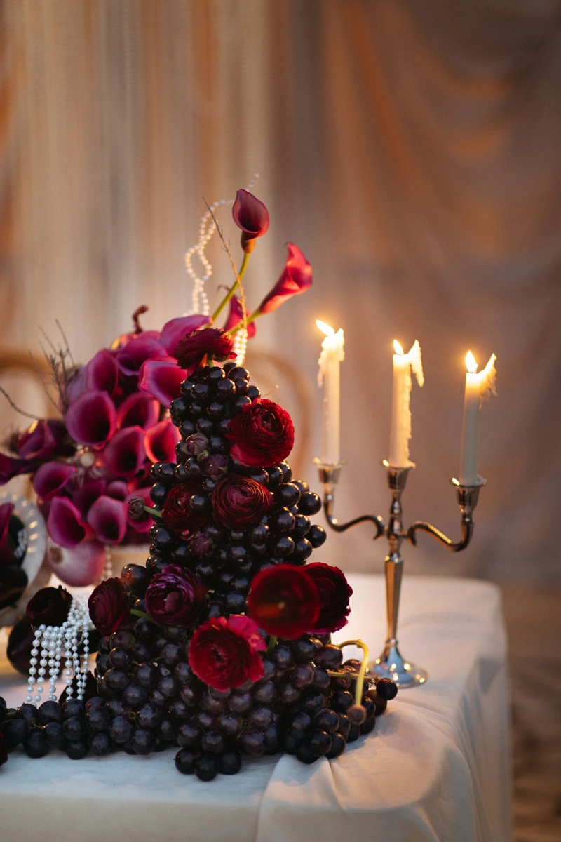 Flottie Shades of Burgundy wedding decor and organisation: intimate evening close-up of a candelabra, dark orchids, eggplants, and red onions creating a moody and luxurious aesthetic
