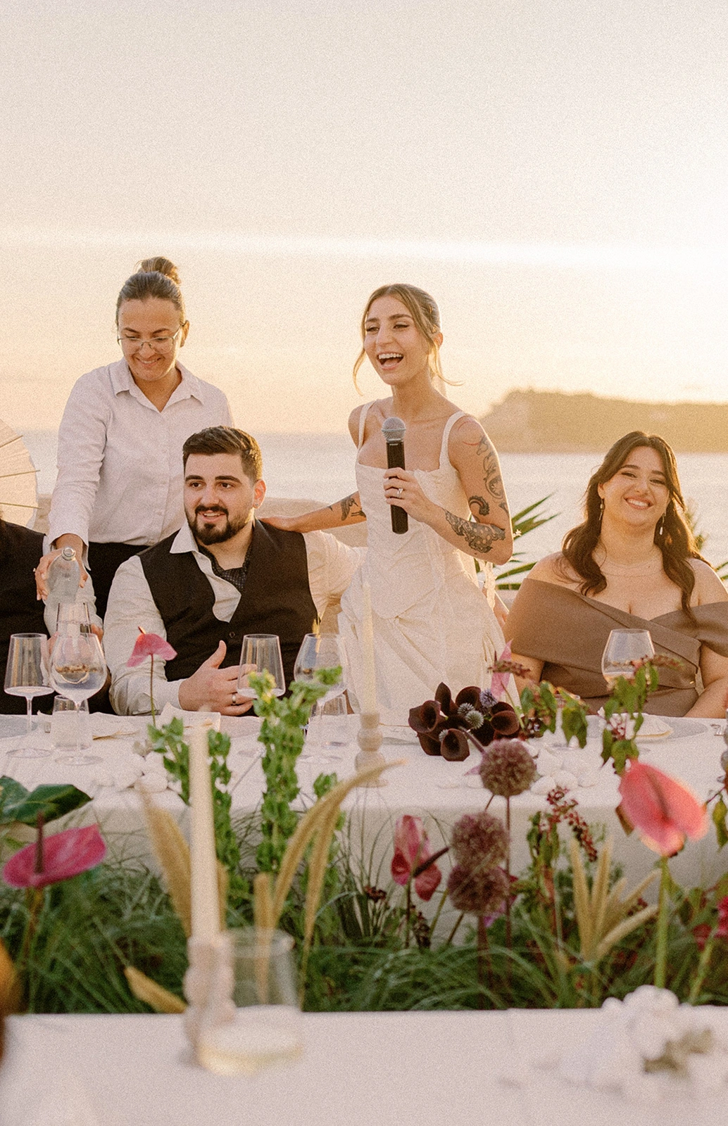 Flottie wedding decor and organisation: joyful bride speaking into a microphone during a wedding reception dinner with guests, candlelit tablescape, and floral arrangements