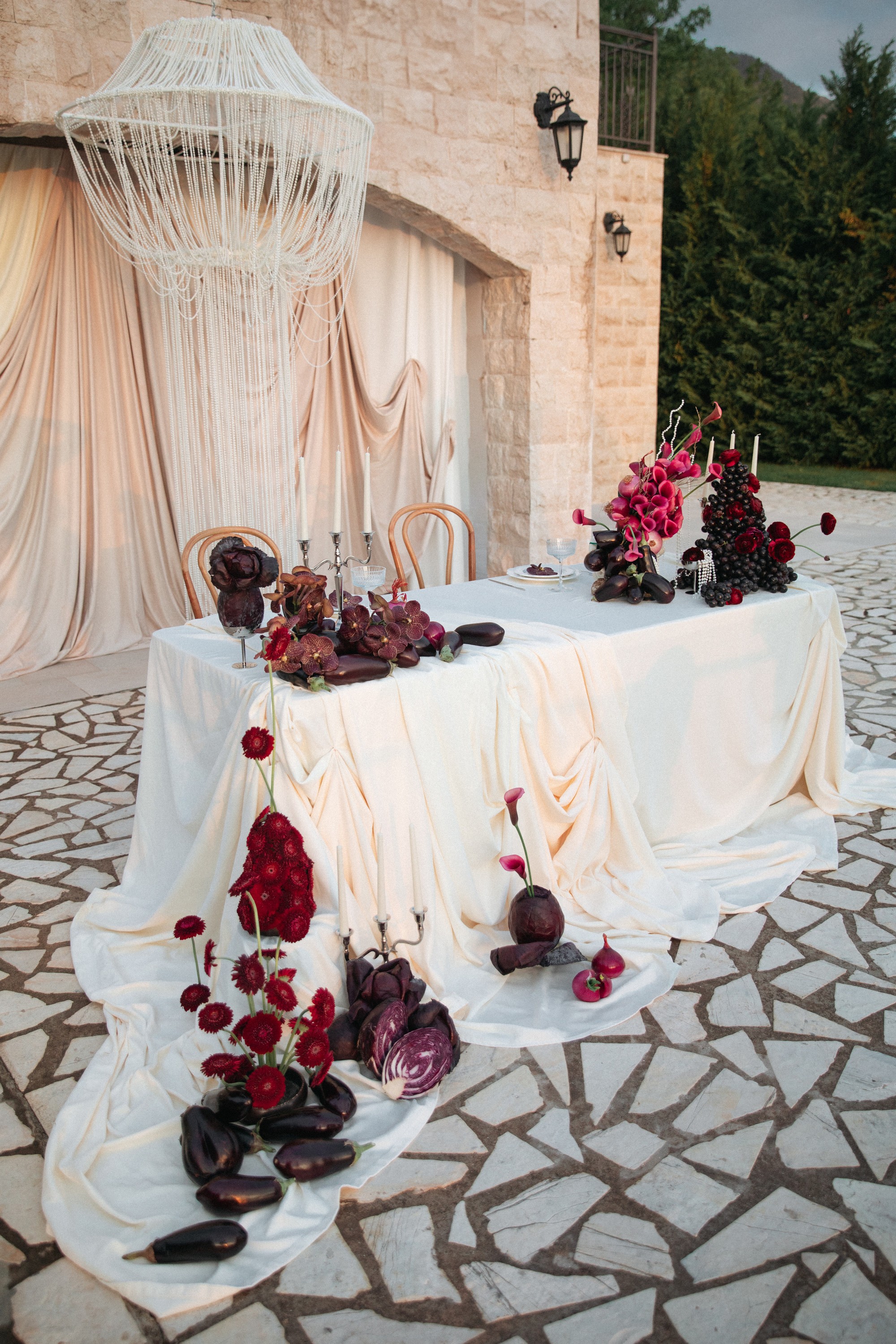 Flottie Shades of Burgundy wedding decor and organisation: close-up of a dramatic tablescape arrangement featuring vibrant red gerberas, fresh eggplants, sliced red cabbage, and a silver candelabra