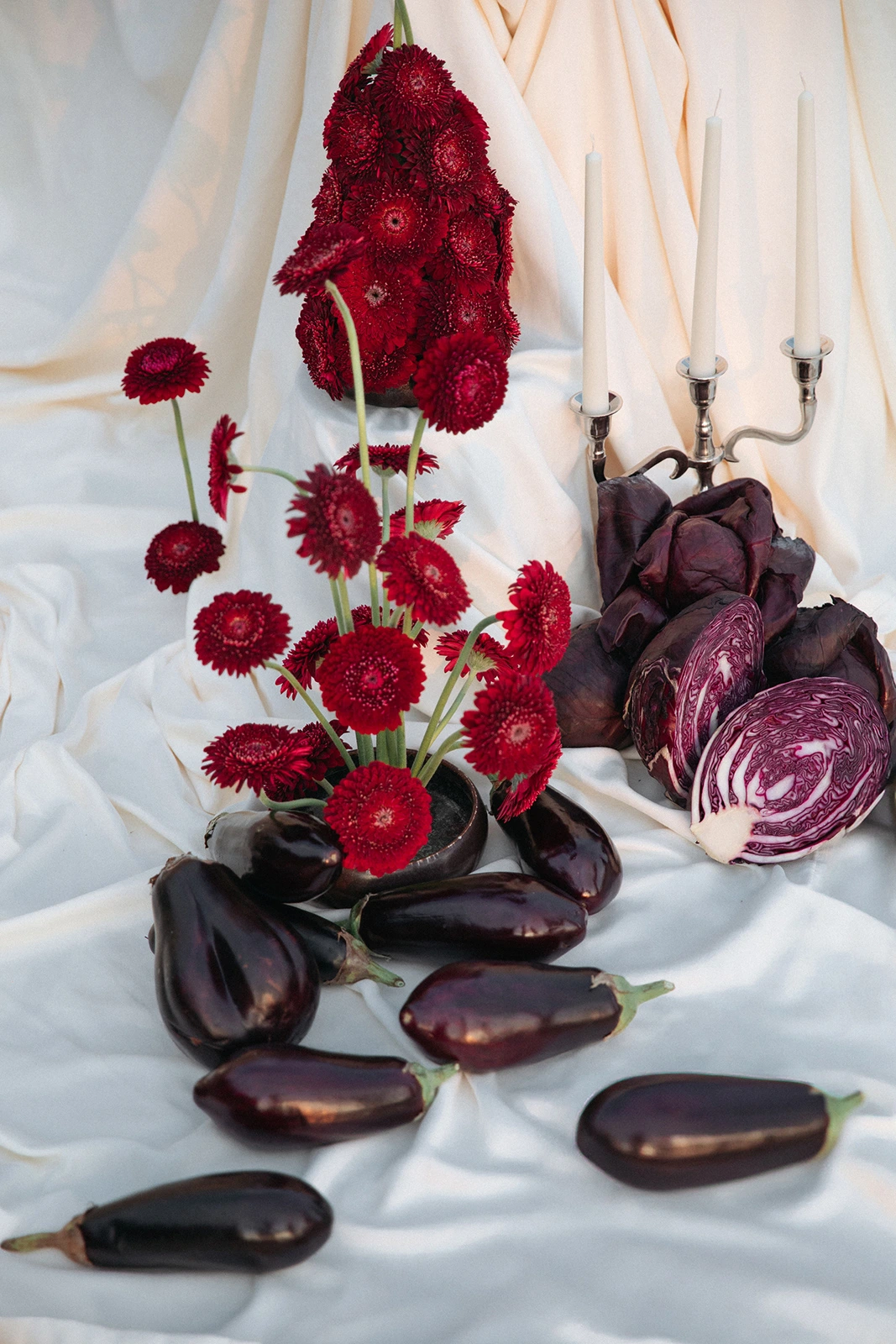 Flottie Shades of Burgundy wedding decor and organisation: close-up of a dramatic tablescape arrangement featuring vibrant red gerberas, fresh eggplants, sliced red cabbage, and a silver candelabra