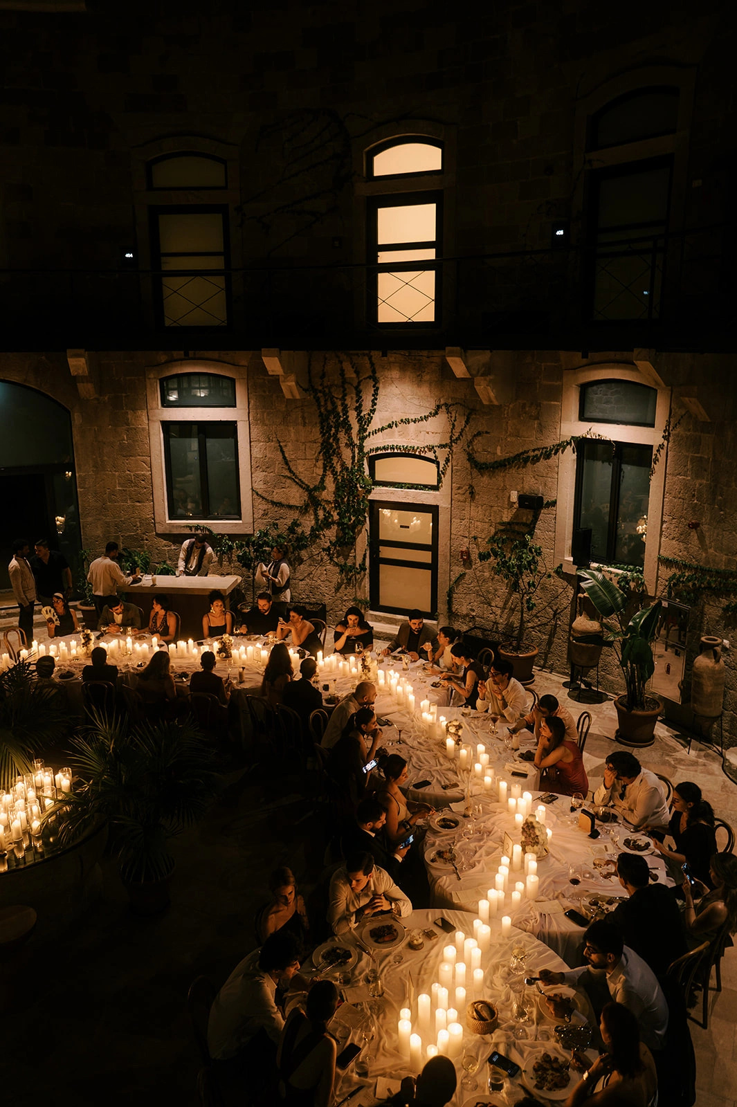 Flottie wedding decor and organisation: high-angle view of an intimate evening wedding reception in a stone courtyard illuminated by numerous candles along long banquet tables