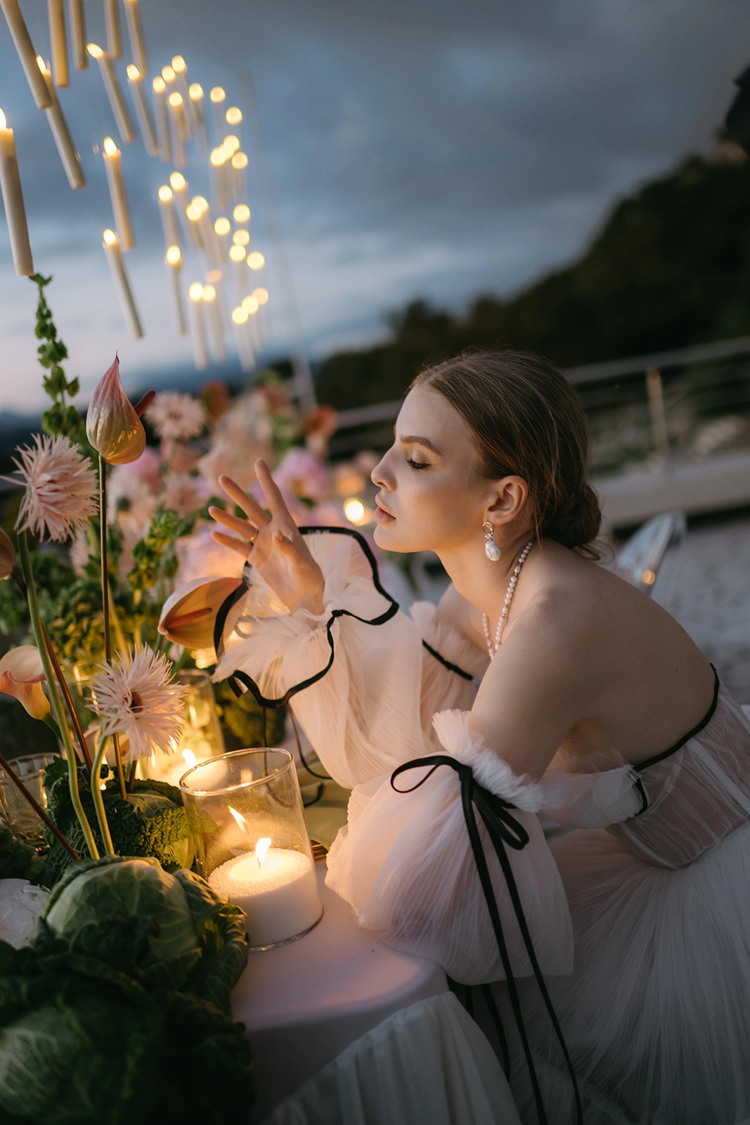 Flottie The Cabbage Affair wedding decor and organisation: close-up portrait of a bride in a tulle dress with black ribbons gently touching exotic anthurium flowers and gerberas at a candlelit evening reception table