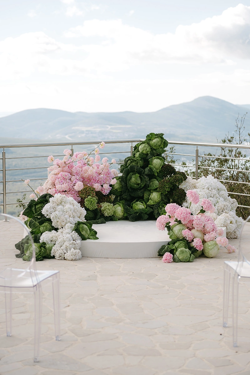 Flottie The Cabbage Affair wedding decor and organisation: modern ceremony setup featuring transparent ghost chairs and a unique sculptural altar installation made of fresh cabbages and pink and white florals overlooking a mountain view