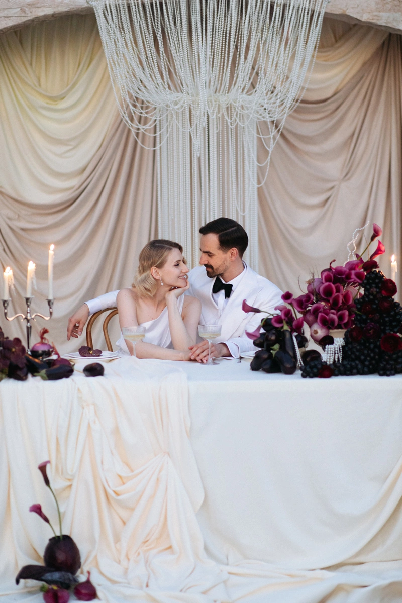 Flottie Shades of Burgundy wedding decor and organisation: bride in a minimalist white gown and groom in a white double-breasted jacket toasting with champagne in front of a draped arch