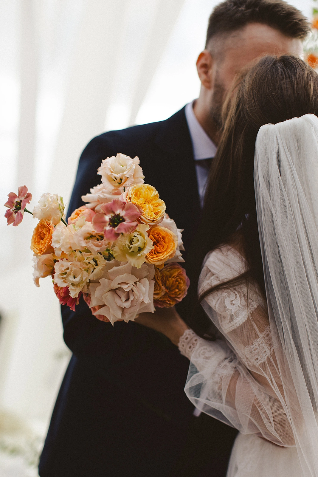 Flottie Peach Haze wedding decor and organisation: bride and groom posing together under a draped archway framed by vibrant peach and pink floral arrangements
