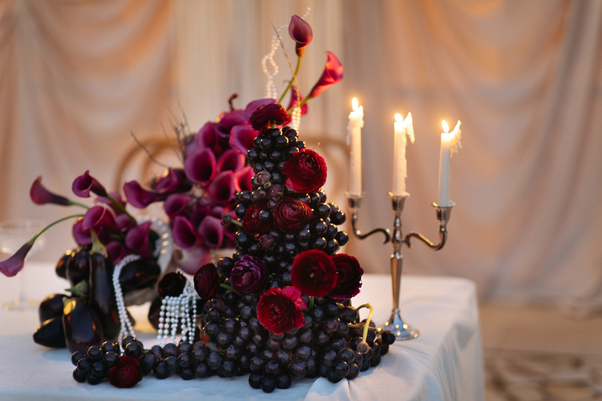 Flottie Shades of Burgundy wedding decor and organisation: intimate evening close-up of a candelabra, dark orchids, eggplants, and red onions creating a moody and luxurious aesthetic