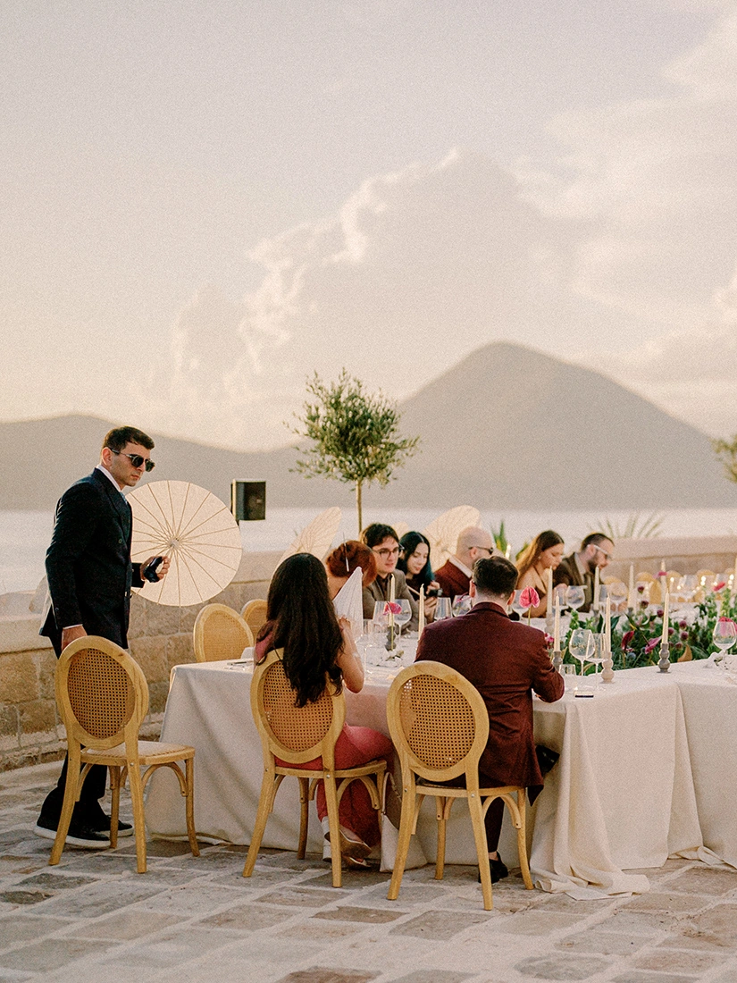 Flottie wedding decor and organisation: scenic outdoor wedding banquet setup by the water with guests dining at elegantly draped tables under a mountain view landscape