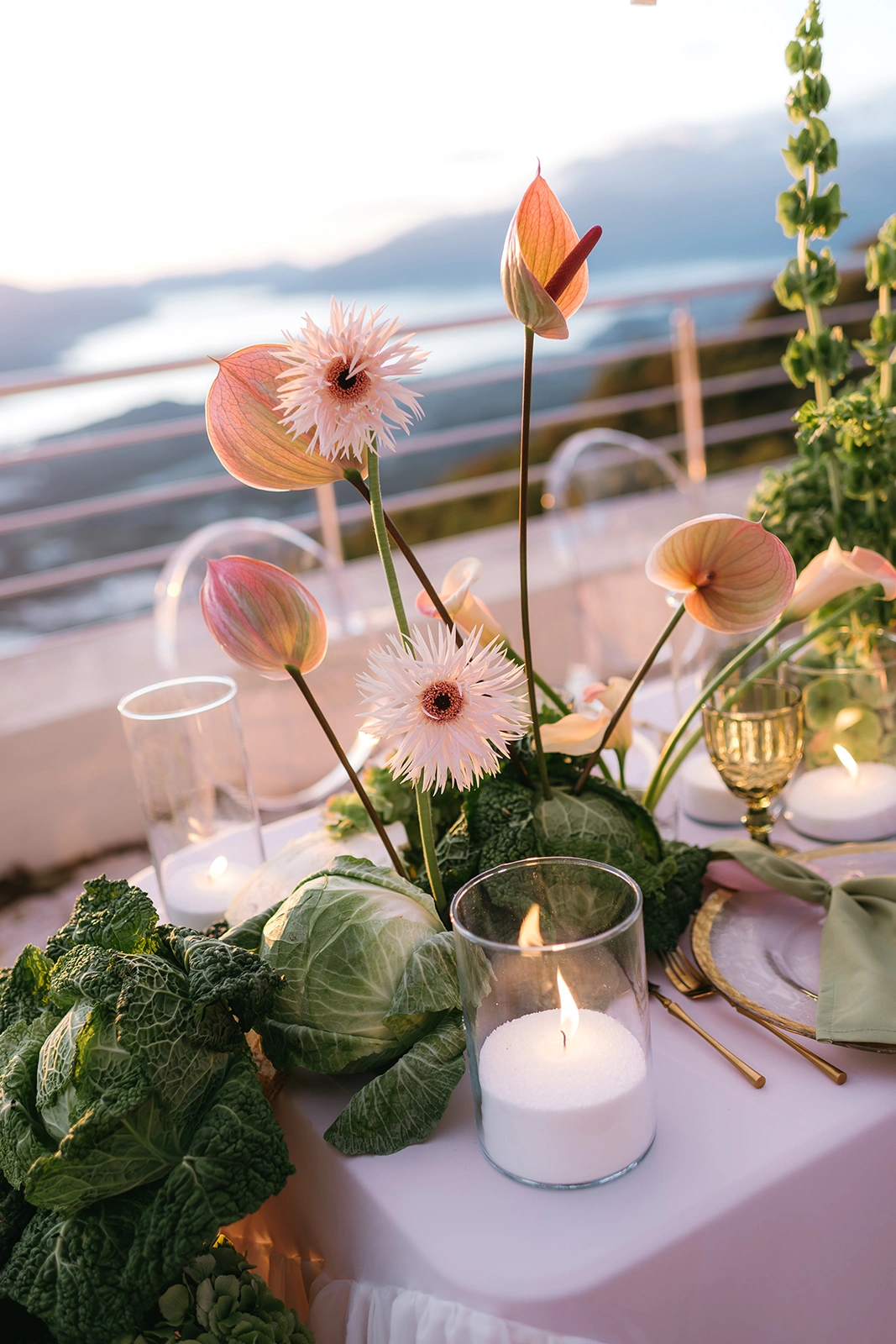 Flottie The Cabbage Affair wedding decor and organisation: close-up of an upscale reception table center-piece featuring fresh heads of cabbage, pastel anthuriums, gerberas, and glowing candles in glass cylinders