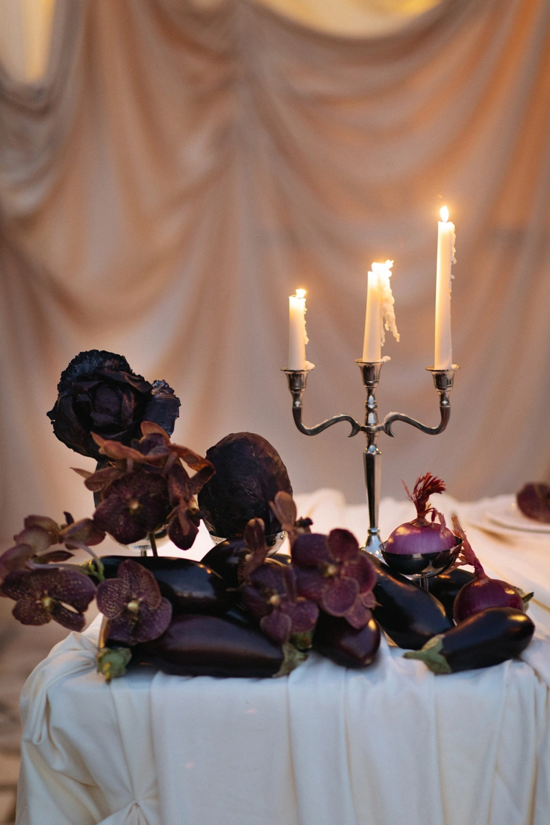 Flottie Shades of Burgundy wedding decor and organisation: intimate evening close-up of a candelabra, dark orchids, eggplants, and red onions creating a moody and luxurious aesthetic