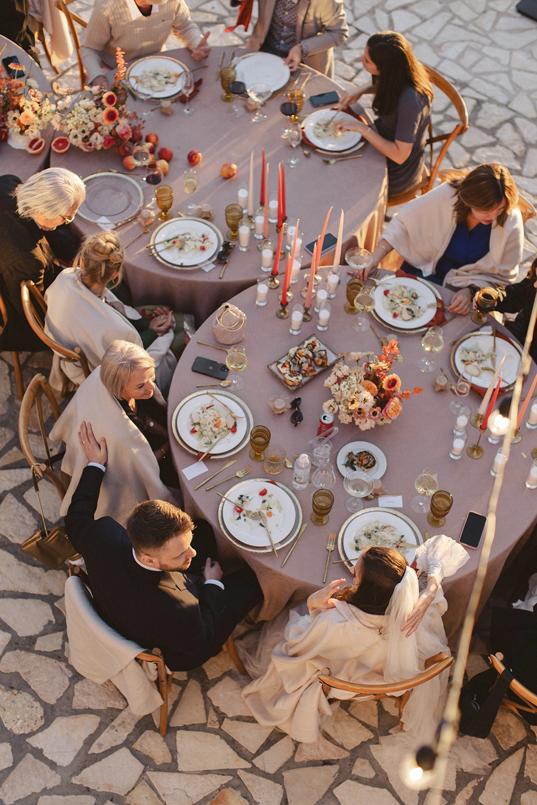 Flottie Peach Haze wedding decor and organisation: aerial view of guests enjoying an outdoor wedding reception dinner at round tables styled with peach-toned florals and candles