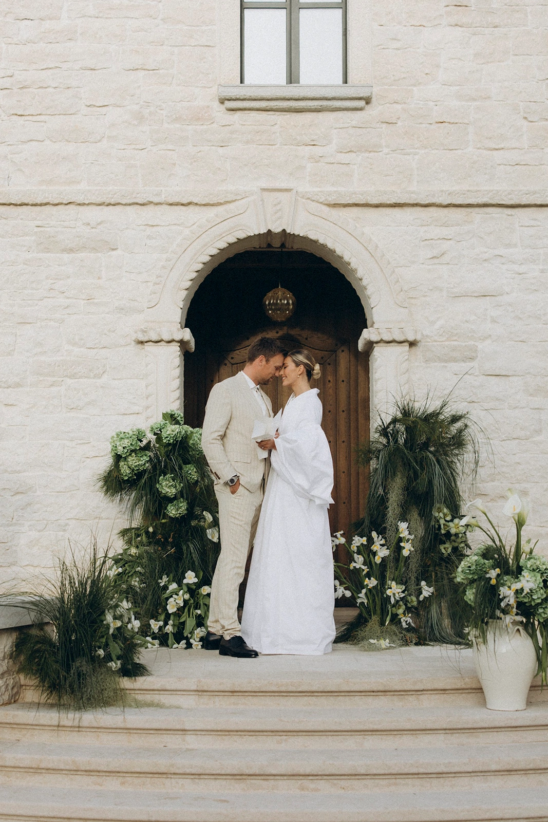 Flottie wedding decor and organisation: bride and groom standing closely together at a rustic stone entrance framed by lush green botanical and floral installations